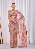 A woman models a light pink and beige floral saree with a beige sleeveless blouse, standing in front of a white curtain backdrop.