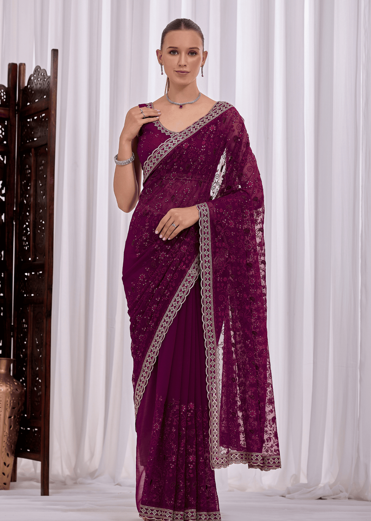 A woman models a deep purple georgette saree with intricate silver floral embroidery and a matching silver border, set against a background of white drapes.