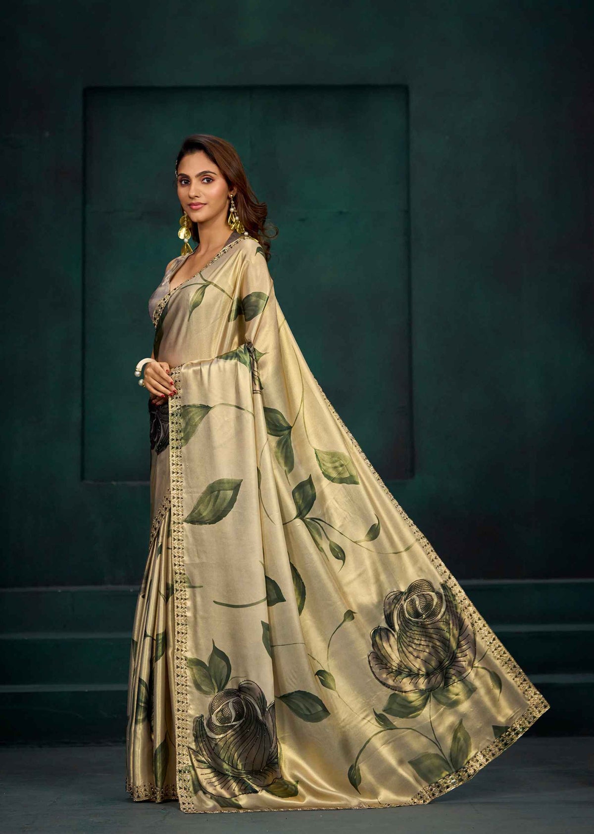 A woman models a beige satin saree with a green rose and leaf print, standing against a dark green textured studio backdrop.