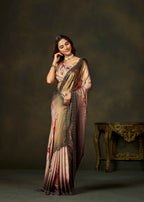 A woman modeling a draped satin saree with a metallic rose gold and beige base, decorated with red floral motifs and a black patterned border, set against a dark brown studio backdrop with an ornate table.