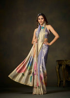 A woman models a draped satin saree with a lavender-purple body, a beige border, and a multicolor floral print on the lower portion. She is posed against a dark brown studio background with a small wooden table visible on the right.