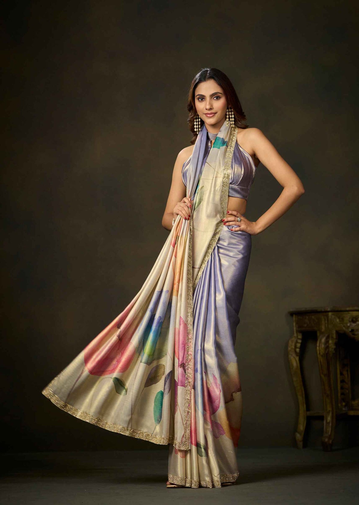 A woman models a draped satin saree with a lavender-purple body, a beige border, and a multicolor floral print on the lower portion. She is posed against a dark brown studio background with a small wooden table visible on the right.