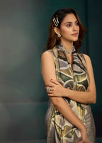 A woman models a sleeveless satin saree with a metallic gold and olive green abstract pattern, paired with a matching dupatta and gold jewelry, set against a dark teal studio background.