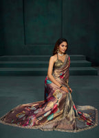 A woman models a draped satin saree with a metallic bronze base and a colorful floral and leaf pattern in pink, teal, and gold, set against a dark teal studio backdrop with steps.