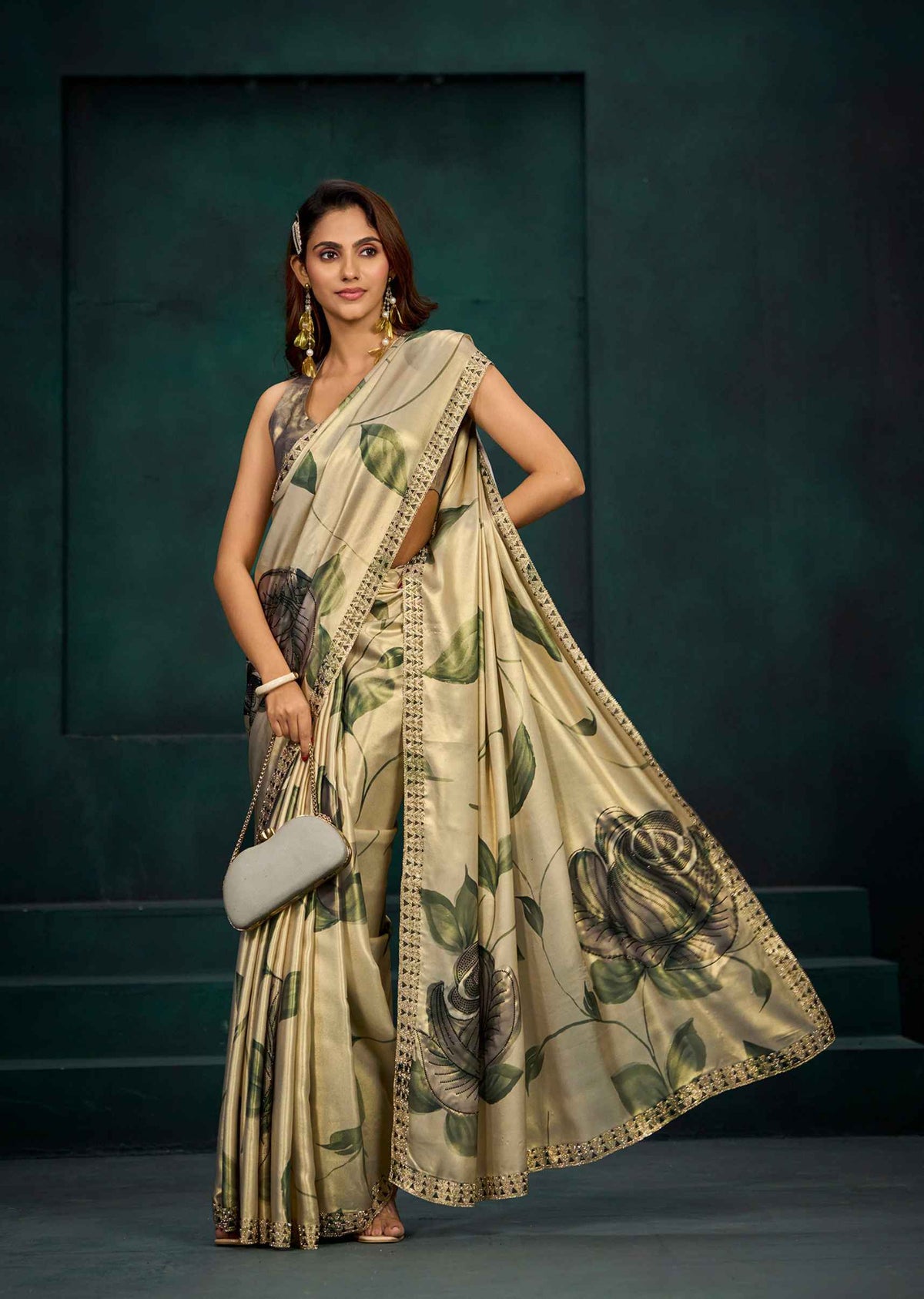 A woman models a beige saree with a green and black floral print, featuring a gold border. She is standing against a dark green textured studio backdrop.
