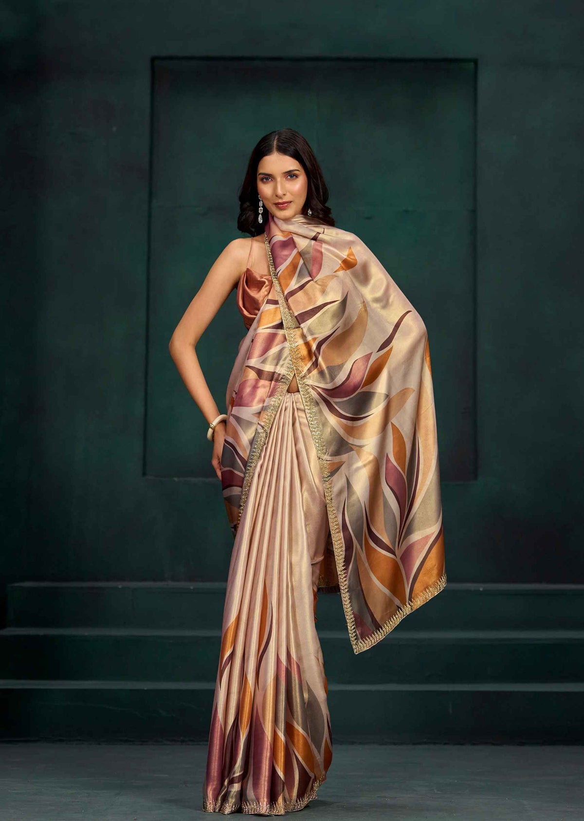 A woman models a draped satin saree with an abstract geometric pattern in beige, brown, orange, and purple tones, set against a dark green studio backdrop.