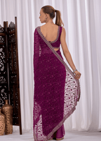A woman shown from the back wearing a deep purple embroidered saree with a matching sleeveless blouse, standing in front of white draped curtains.