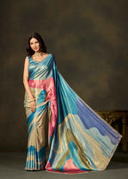 A woman models a silk saree with a multicolor abstract print featuring shades of blue, pink, beige, and gold. The saree has a matching blue embroidered blouse and is posed to show the drape and pallu against a dark, textured studio background.
