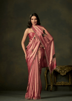 A woman models a metallic pink silk saree with a matching blouse, standing against a dark brown studio backdrop with an ornate gold table visible on the right.