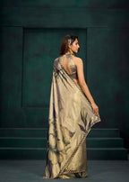 A woman stands with her back to the camera wearing a draped beige saree with a large floral print and a patterned border, posed against a dark green textured studio backdrop.