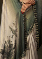 A close-up of a draped traditional garment with a sage green and cream gradient, featuring black and white floral embroidery and a black-and-gold patterned border. A model's hands with red nail polish are visible holding the fabric.