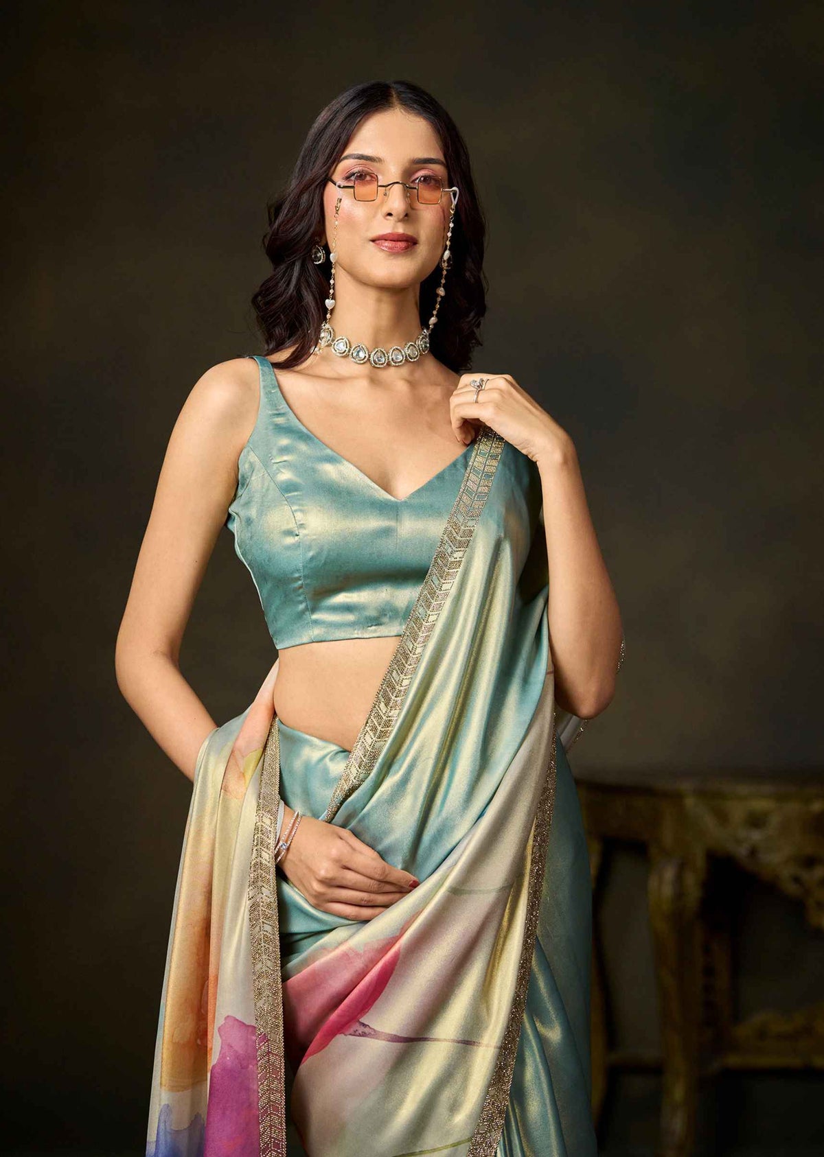A woman models a satin saree with a light blue-green blouse and a matching draped pallu featuring a pastel gradient of pink, yellow, and blue with a gold border. She is wearing glasses, a silver necklace, and earrings, posed against a dark, moody studio background.