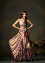 A woman models a floor-length draped gown with a metallic rose-gold and dark olive-green base, featuring a pink and red floral pattern along the lower skirt, set against a dark textured studio backdrop.
