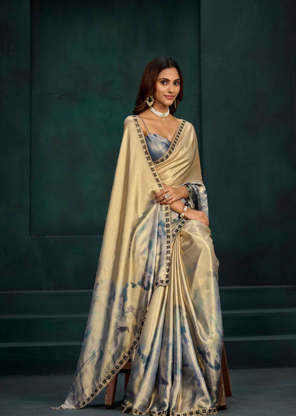 A woman models a beige and blue watercolor-printed silk saree with black border detailing, standing against a dark green studio backdrop.