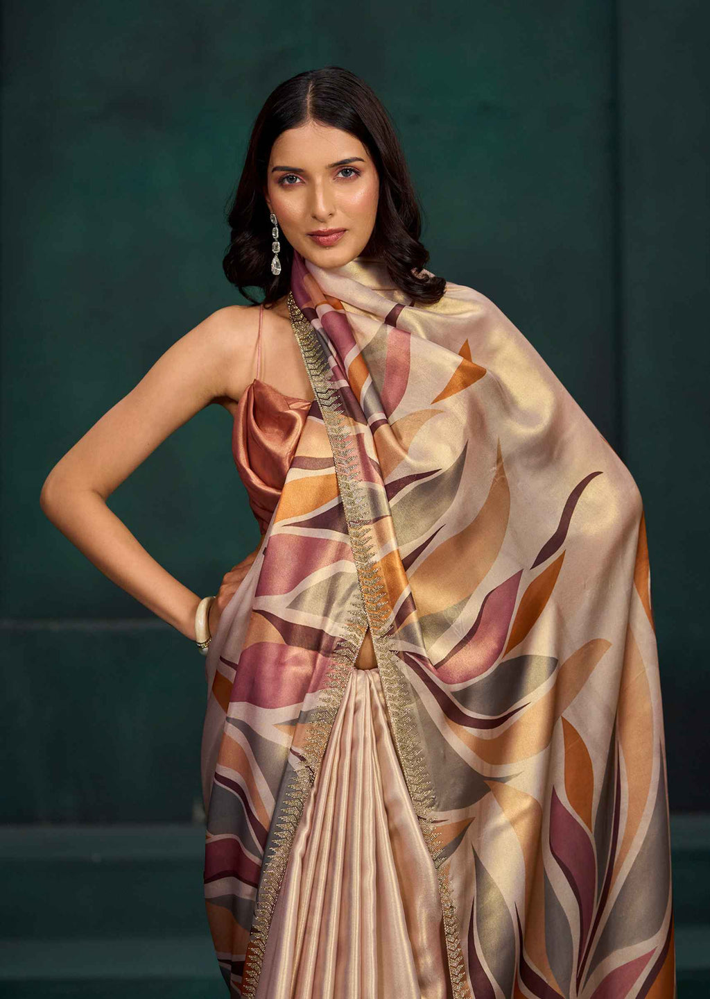 A woman models a draped satin saree with a large matching scarf, featuring an abstract leaf pattern in beige, gold, brown, and muted pink, set against a dark green textured studio background.