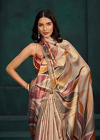 A woman models a draped satin saree with a large matching scarf, featuring an abstract leaf pattern in beige, gold, brown, and muted pink, set against a dark green textured studio background.