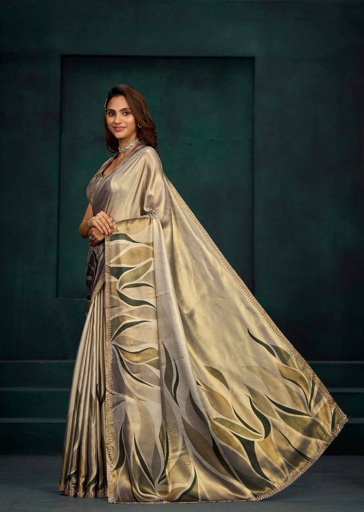 A woman modeling a metallic champagne-gold saree with a large draped pallu featuring dark olive-green leaf-like patterns, posed against a dark teal studio backdrop.