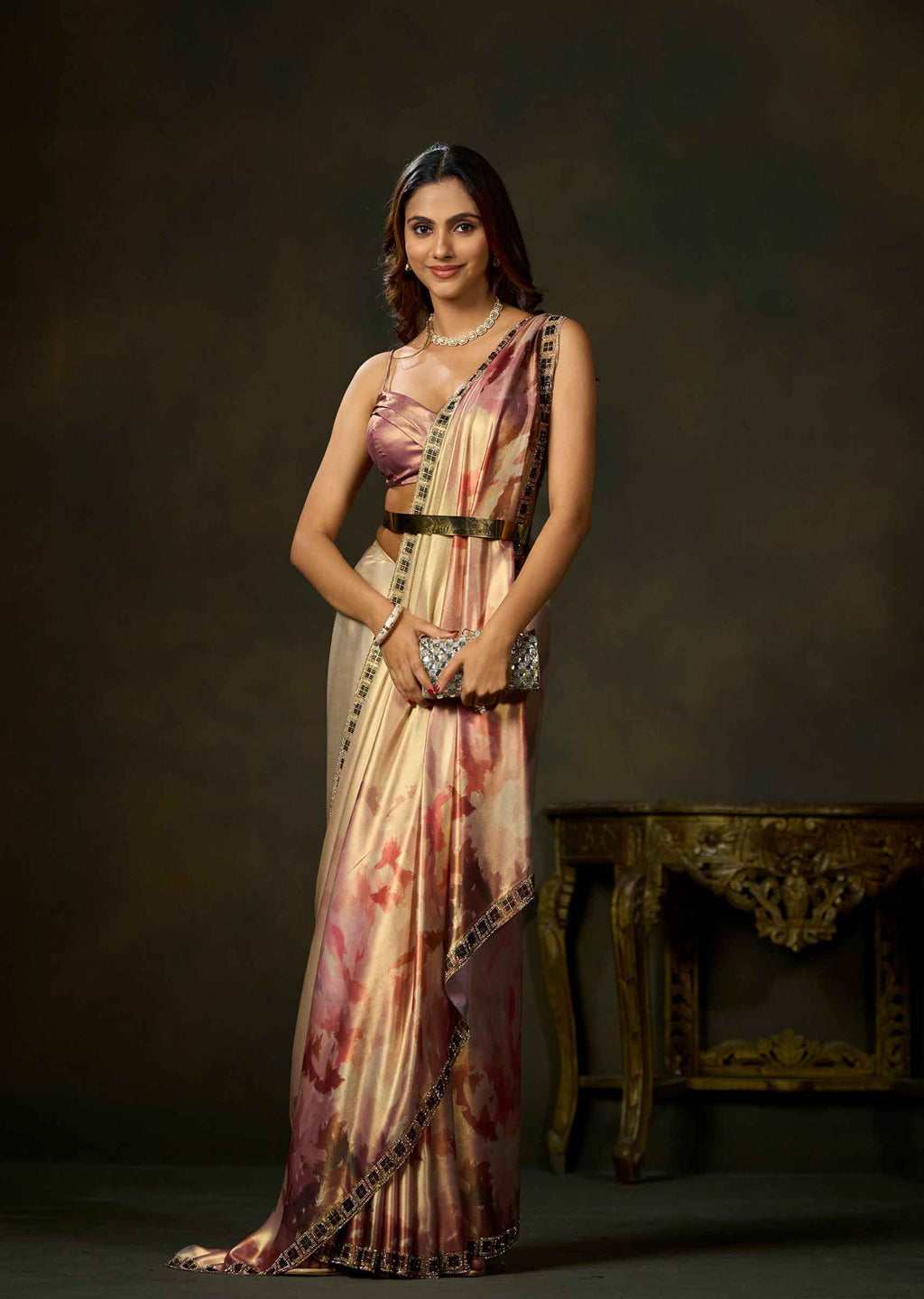 A woman models a traditional Indian saree with a pink and beige floral print, paired with a matching crop top and belt, standing in front of a dark studio background.