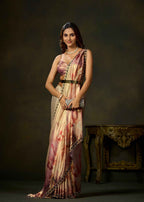 A woman models a traditional Indian saree with a pink and beige floral print, paired with a matching crop top and belt, standing in front of a dark studio background.
