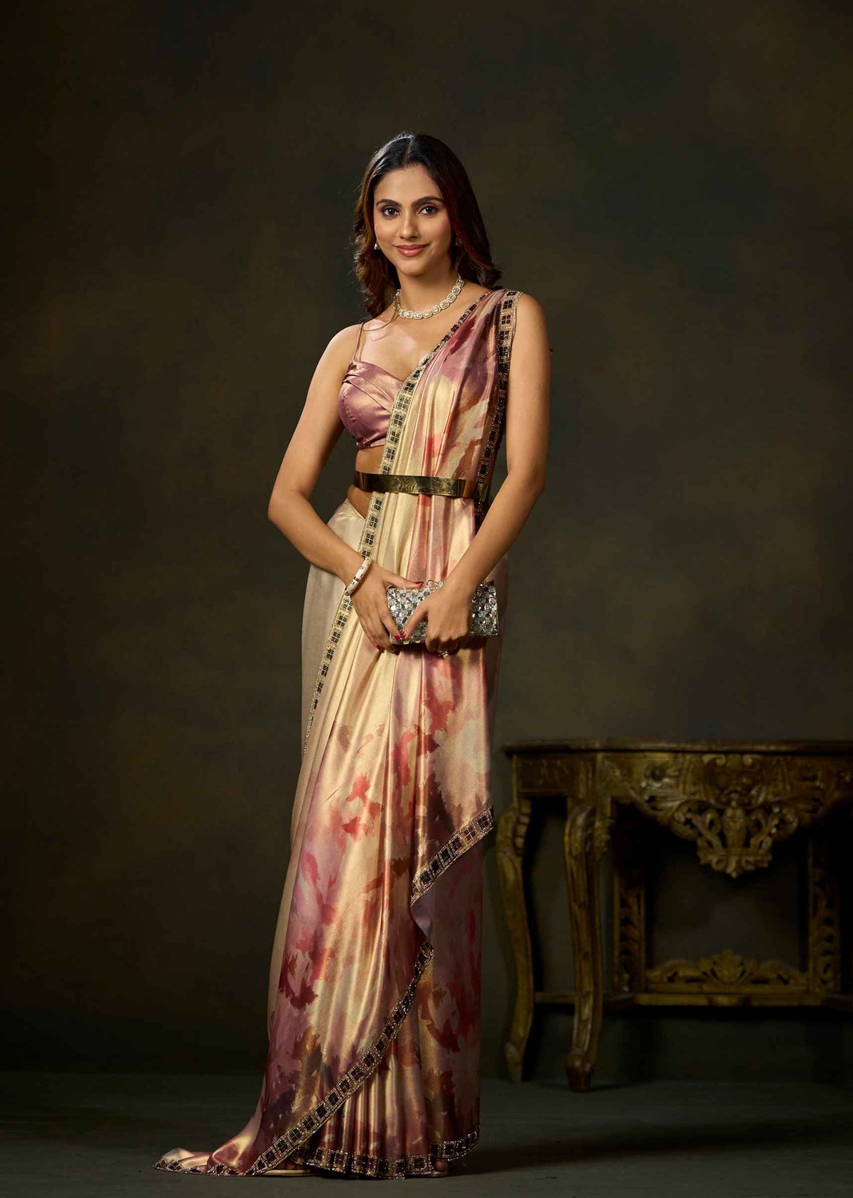 A woman models a traditional Indian saree with a pink and beige floral print, paired with a matching crop top and belt, standing in front of a dark studio background.