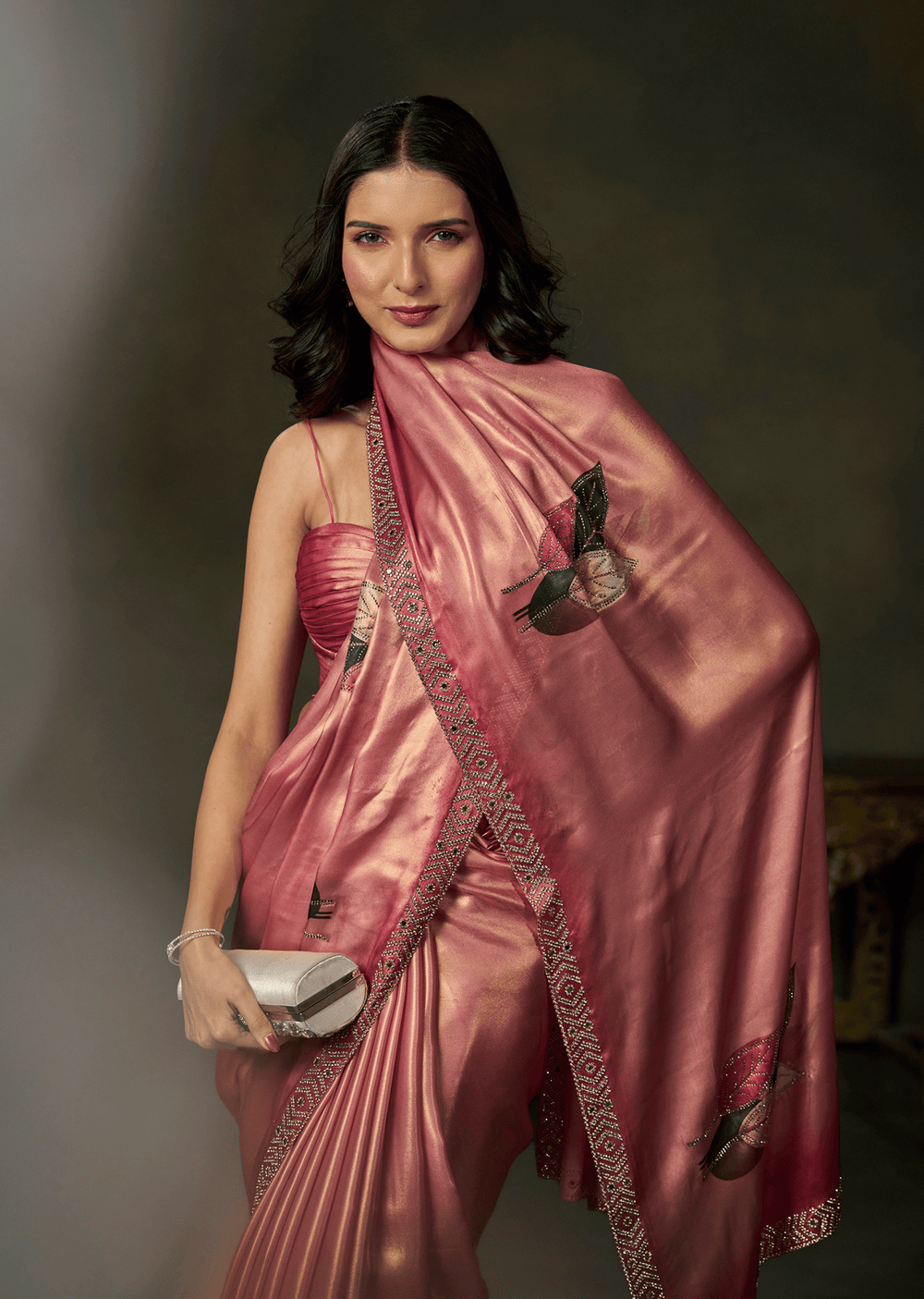 A woman modeling a metallic pink sari with embroidered details and a matching pink strapless top, holding a silver clutch against a dark studio background.
