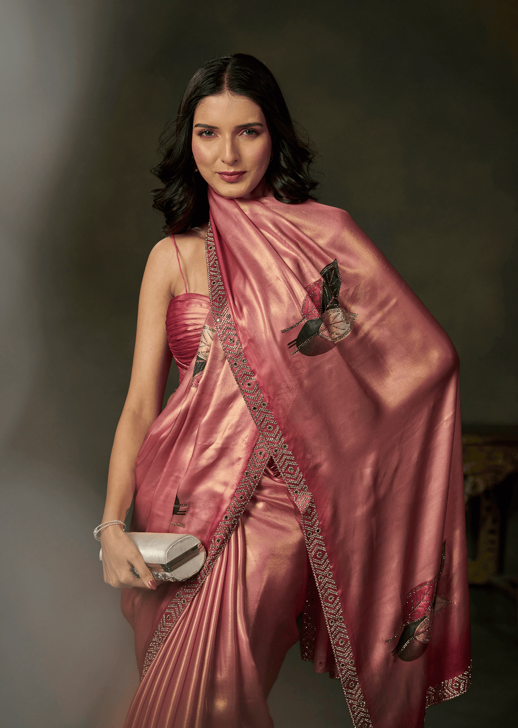 A woman modeling a metallic pink sari with embroidered details and a matching pink strapless top, holding a silver clutch against a dark studio background.