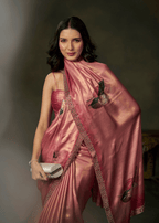 A woman modeling a metallic pink sari with embroidered details and a matching pink strapless top, holding a silver clutch against a dark studio background.