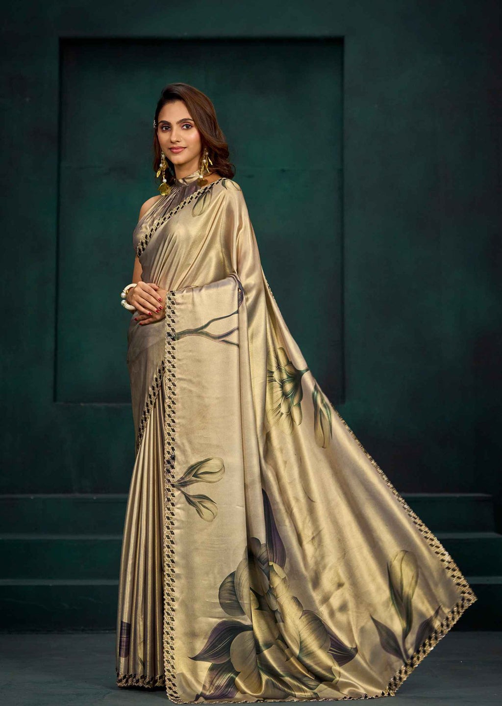 A woman models a champagne beige silk saree with a dark green and black floral print, paired with a matching pleated skirt and gold jewelry, set against a dark green textured studio backdrop.
