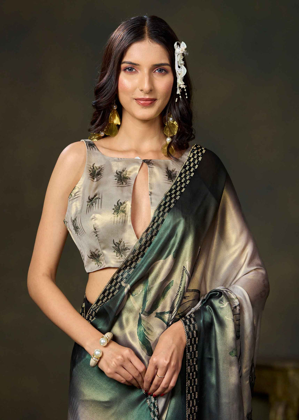 A woman modeling a traditional Indian saree with a metallic green and gold body, a printed beige blouse, and a black and gold border, set against a dark studio background.