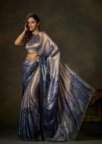 A woman models a metallic lavender-blue saree with a matching crop top against a dark brown studio backdrop. The fabric features a shimmering satin-like sheen and a blue floral vine print along the border.