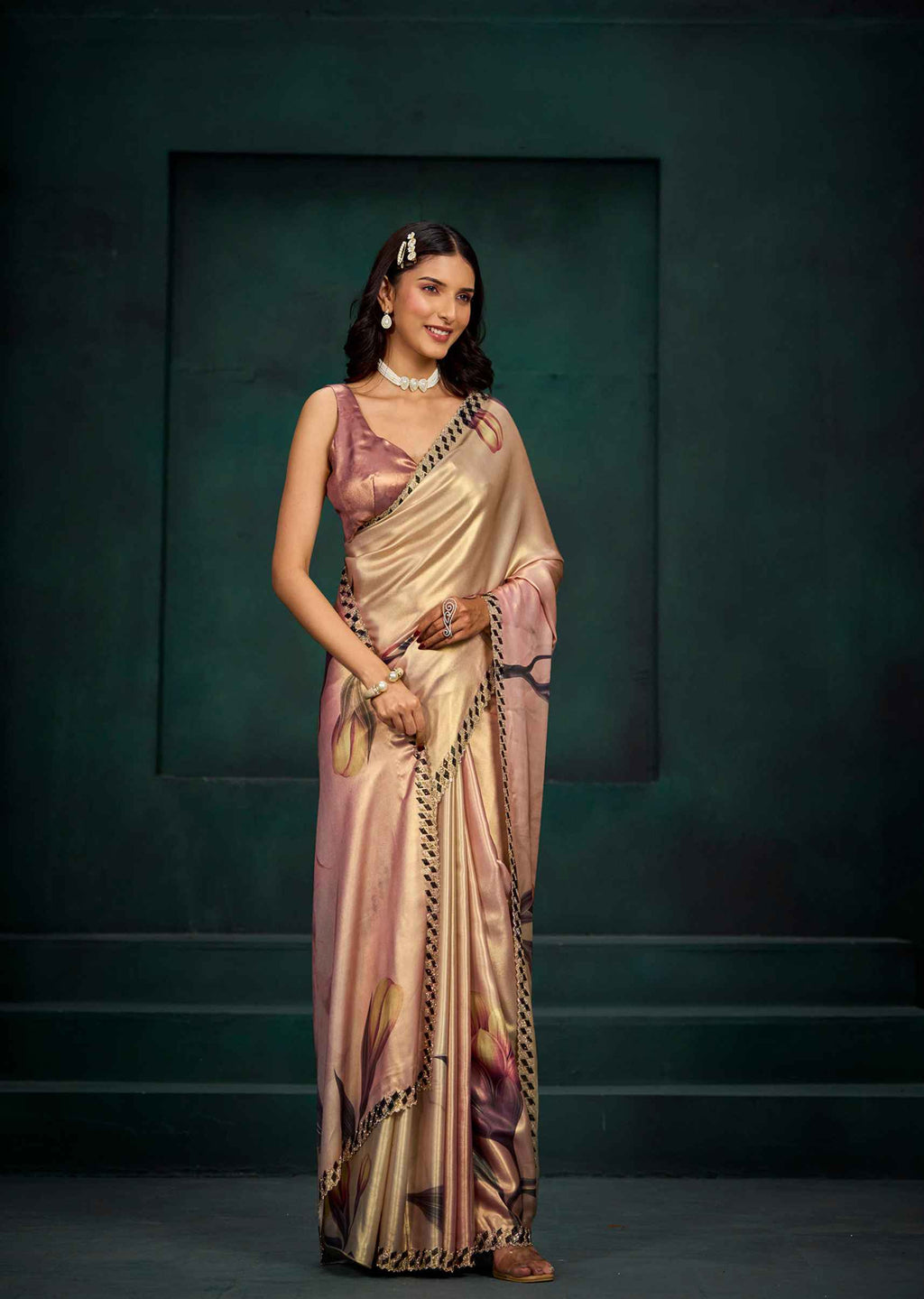 A woman modeling a draped satin saree with a metallic gold base, pink floral patterns, and a black-and-gold border, set against a dark teal studio backdrop.