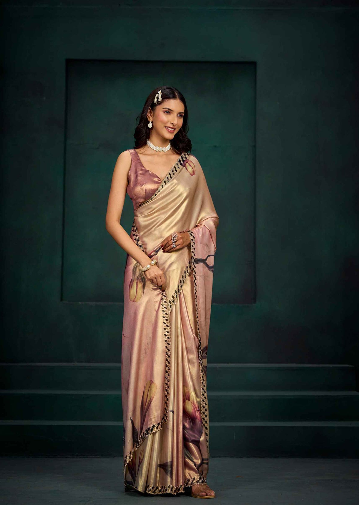 A woman modeling a draped satin saree with a metallic gold base, pink floral patterns, and a black-and-gold border, set against a dark teal studio backdrop.