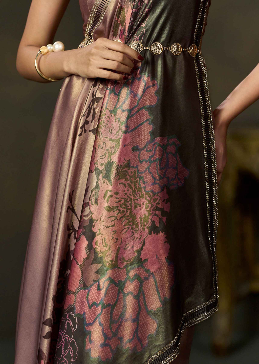A close-up of a woman wearing a shimmering satin dress with a large pink floral brocade pattern over a dark base, styled with a gold chain belt and pearl bracelets.