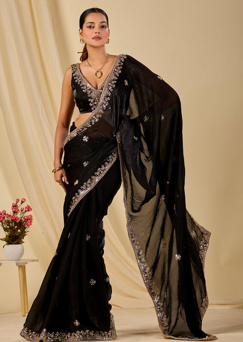 A woman models a black sheer saree with silver embroidered borders and floral motifs, paired with a matching black blouse, against a warm beige draped backdrop.