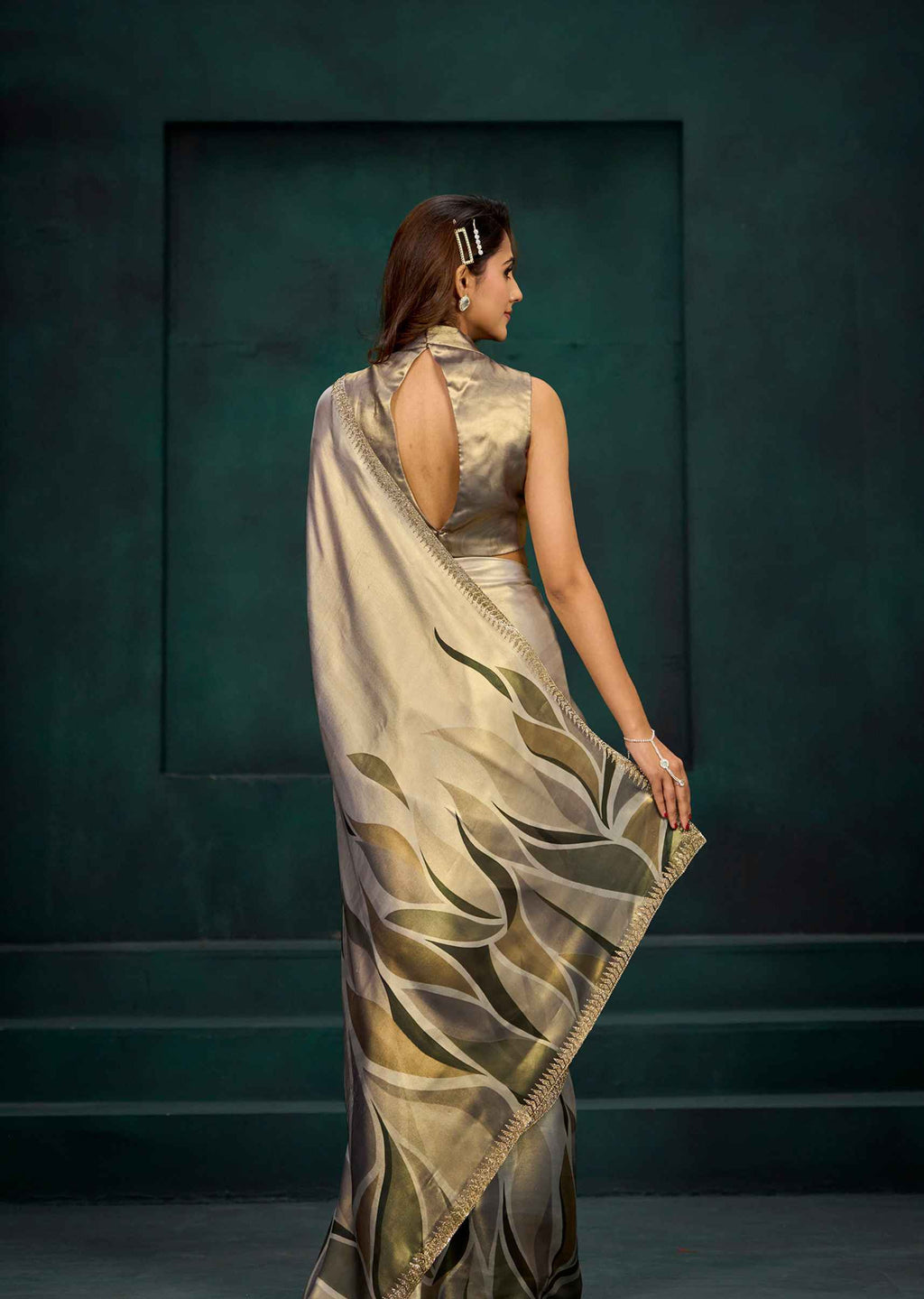 A woman wearing a sleeveless beige saree with a metallic gold and olive green leaf pattern, shown from the back against a dark teal studio backdrop.