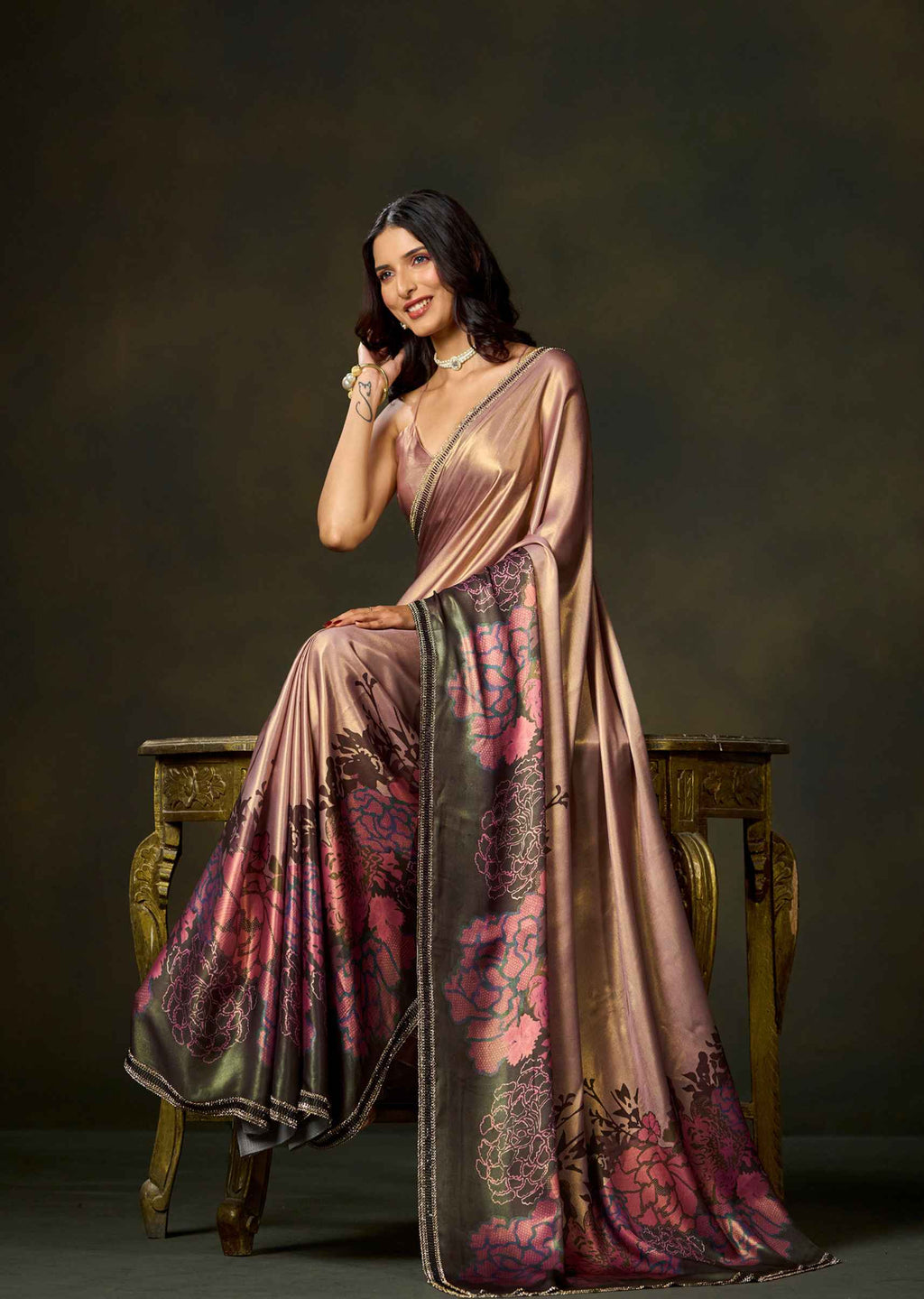A woman seated on an ornate wooden stool wearing a draped satin saree with a metallic bronze-pink base and large floral motifs in pink, burgundy, and dark green. The backdrop is a dark studio background.
