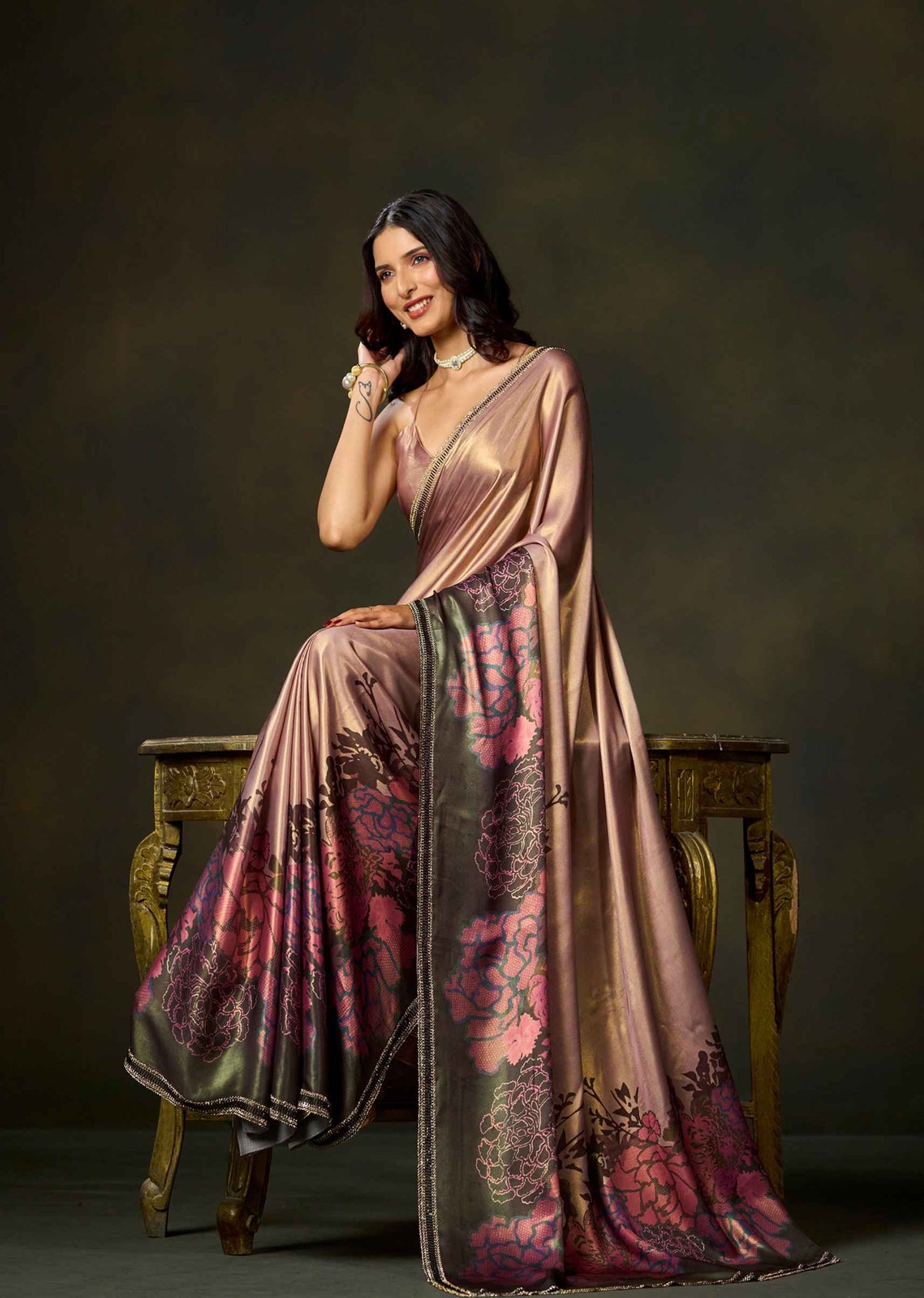 A woman seated on an ornate wooden stool wearing a draped satin saree with a metallic bronze-pink base and large floral motifs in pink, burgundy, and dark green. The backdrop is a dark studio background.