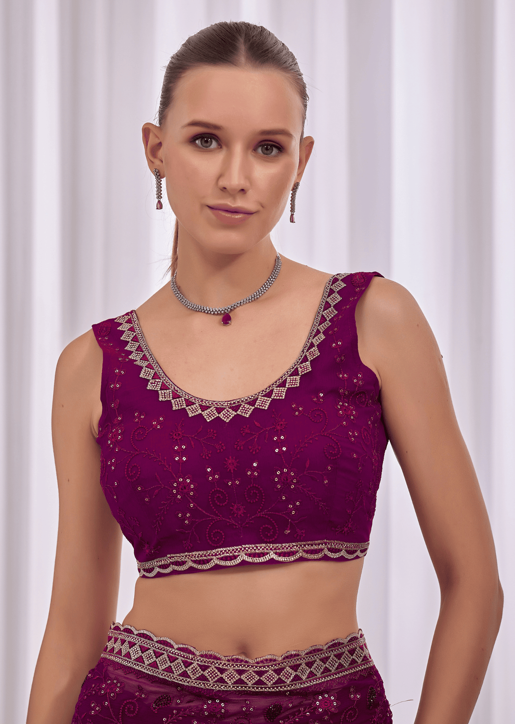 A woman modeling a deep purple embroidered sleeveless blouse with silver mirror work and a matching waistband, set against a soft white curtain background.