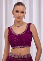 A woman modeling a deep purple embroidered sleeveless blouse with silver mirror work and a matching waistband, set against a soft white curtain background.
