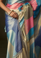 A close-up of a draped saree with a multicolor abstract print in shades of blue, pink, beige, and gray, featuring a gold zari border and a hand wearing a pearl bracelet.