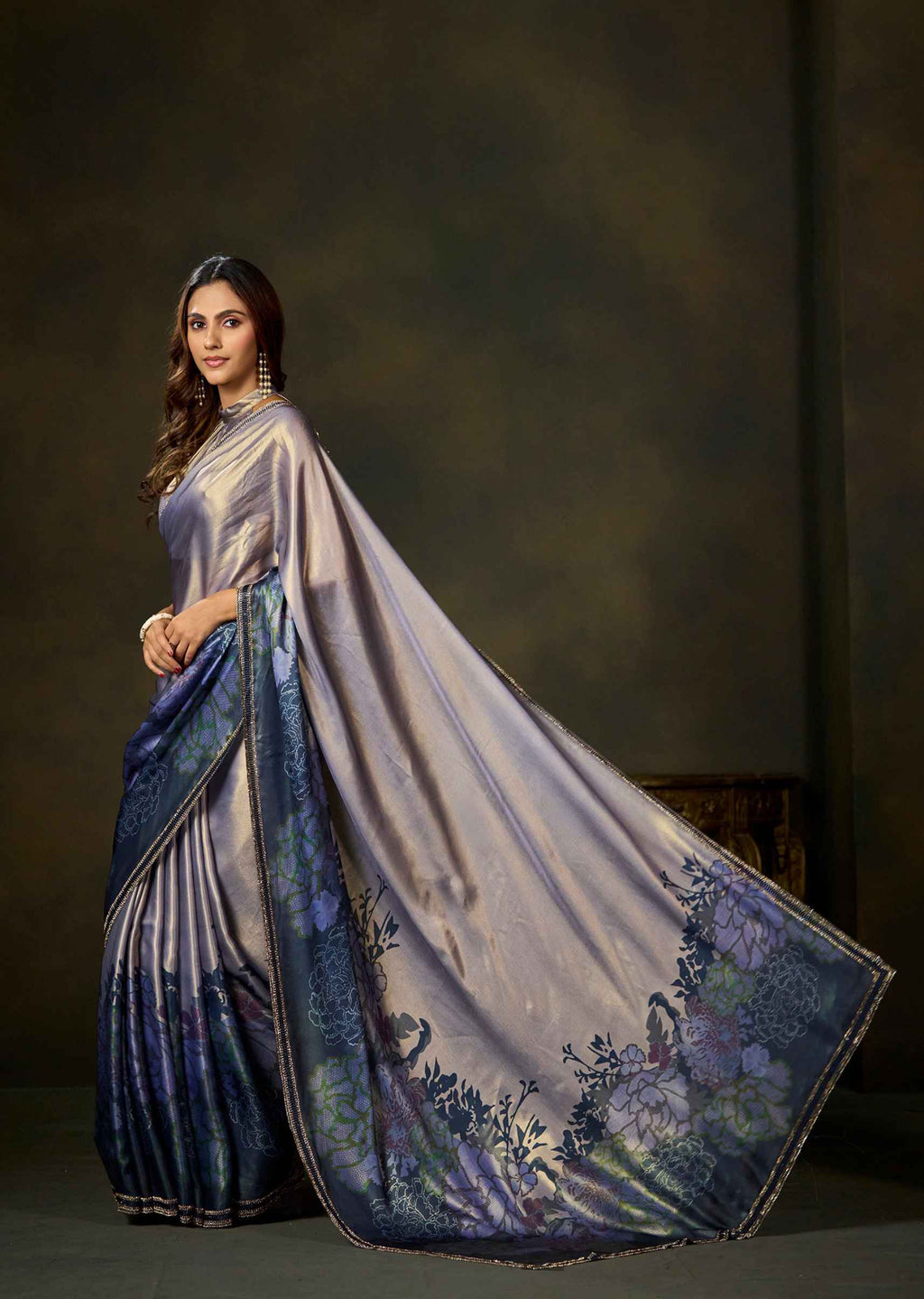 A woman modeling a draped saree with a shimmering lavender-gray body and a dark blue border featuring floral and peacock motifs, set against a dark textured studio backdrop.