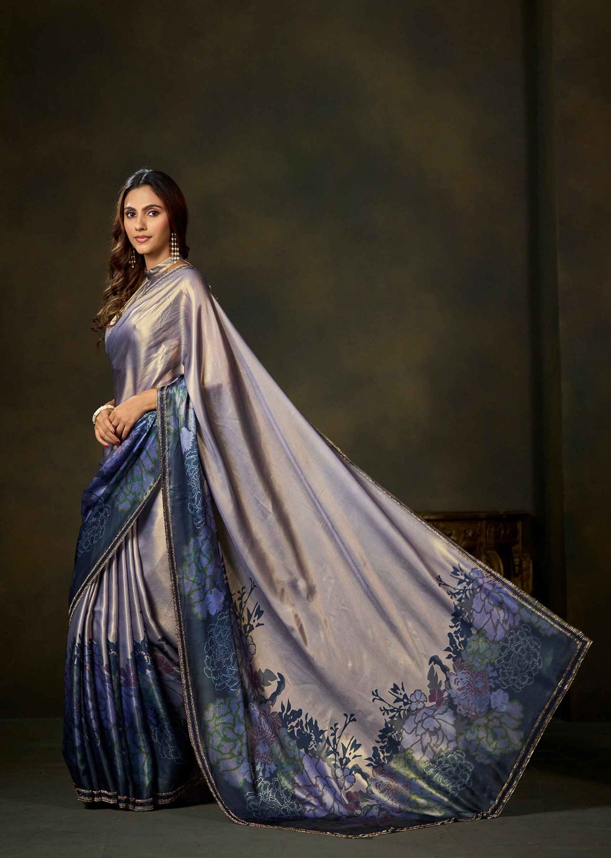 A woman modeling a draped saree with a shimmering lavender-gray body and a dark blue border featuring floral and peacock motifs, set against a dark textured studio backdrop.