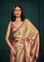 A woman modeling a shiny champagne-gold saree with a pinkish satin blouse, featuring a floral print on the pallu and a black and gold border, set against a dark green background.