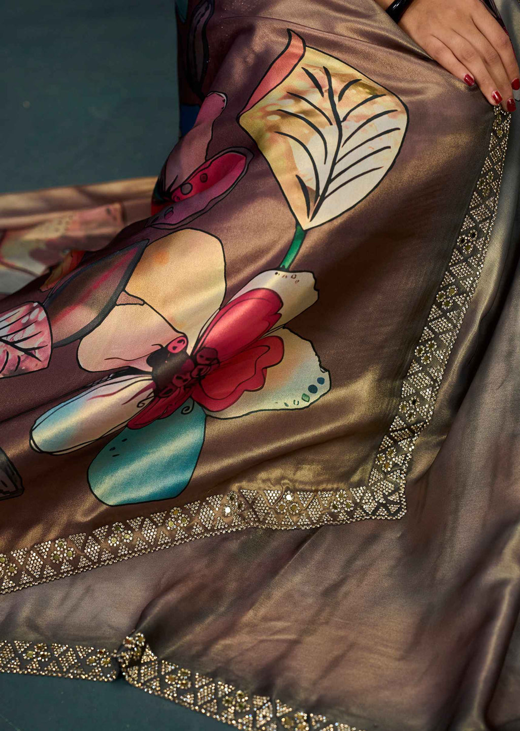 A close-up of a brown satin fabric, likely a sari, featuring a large colorful floral print in red, beige, and teal, finished with an ornate gold embroidered border. A hand with red painted nails is visible holding the fabric.