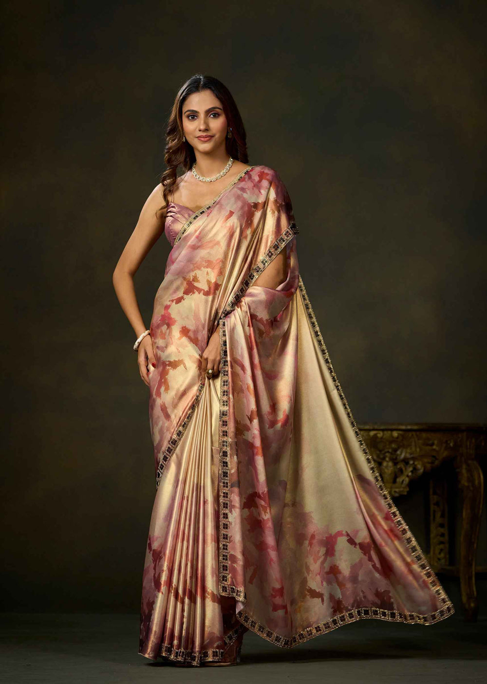 A woman modeling a draped satin saree with a pink and beige floral pattern and a black embroidered border, set against a dark brown studio background.
