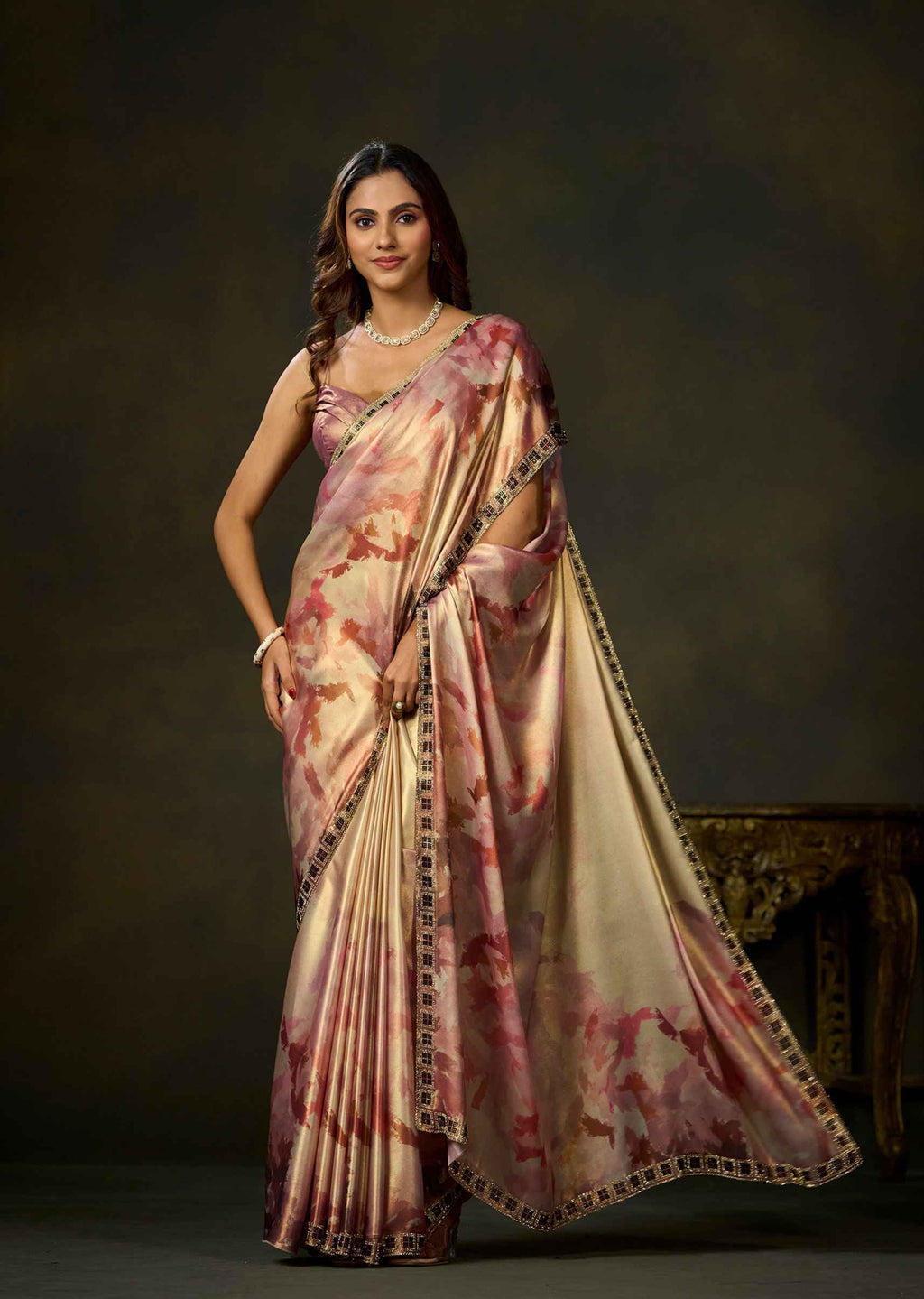 A woman modeling a draped satin saree with a pink and beige floral pattern and a black embroidered border, set against a dark brown studio background.