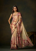 A woman modeling a draped satin saree with a pink and beige floral pattern and a black embroidered border, set against a dark brown studio background.
