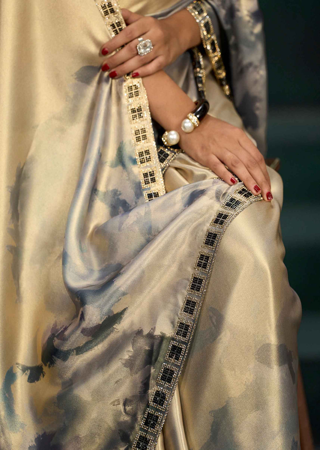 A close-up of a woman wearing a beige and blue watercolor-print saree with black and gold border detailing, accessorized with a pearl and gold bracelet and a large ring.