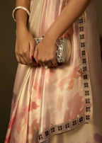 A close-up of a person wearing a light pink satin dress with a watercolor-like darker pink pattern and a decorative black-and-beige checkered border. The person is holding a silver embellished clutch and wearing a white beaded bracelet.