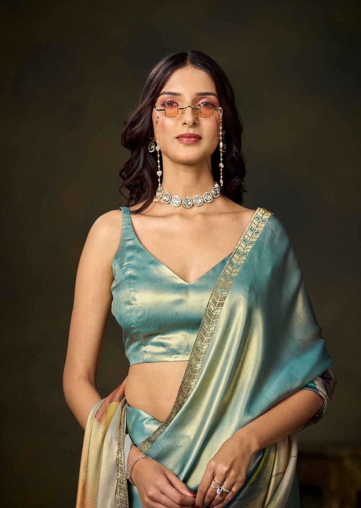 A woman wearing a shimmering teal-green satin saree with gold zari embroidery and a matching sleeveless blouse, styled with statement jewelry against a dark studio background.